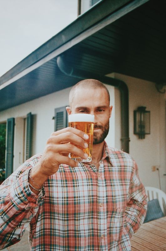 Un homme en chemise à carreaux lève un verre de bière devant la maison