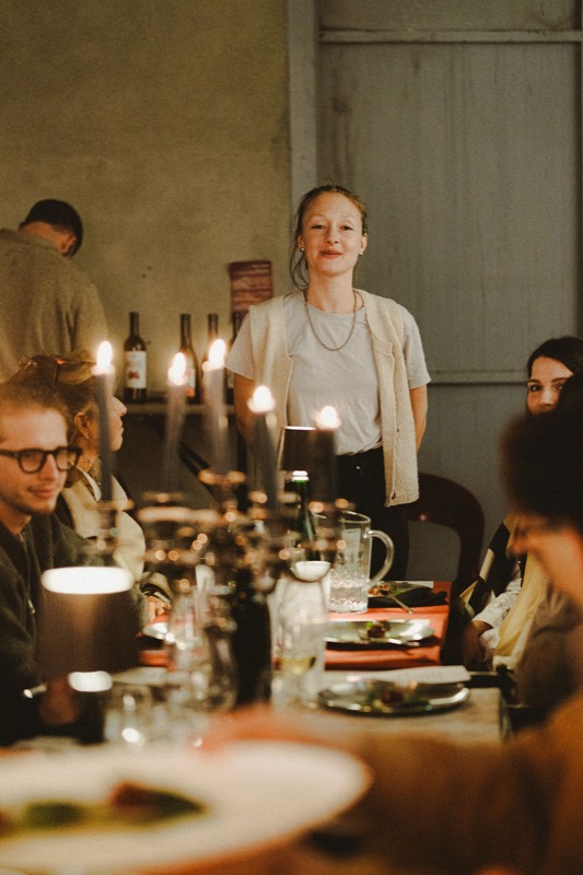 Une femme debout sourit au bout de la table, les convives assis autour